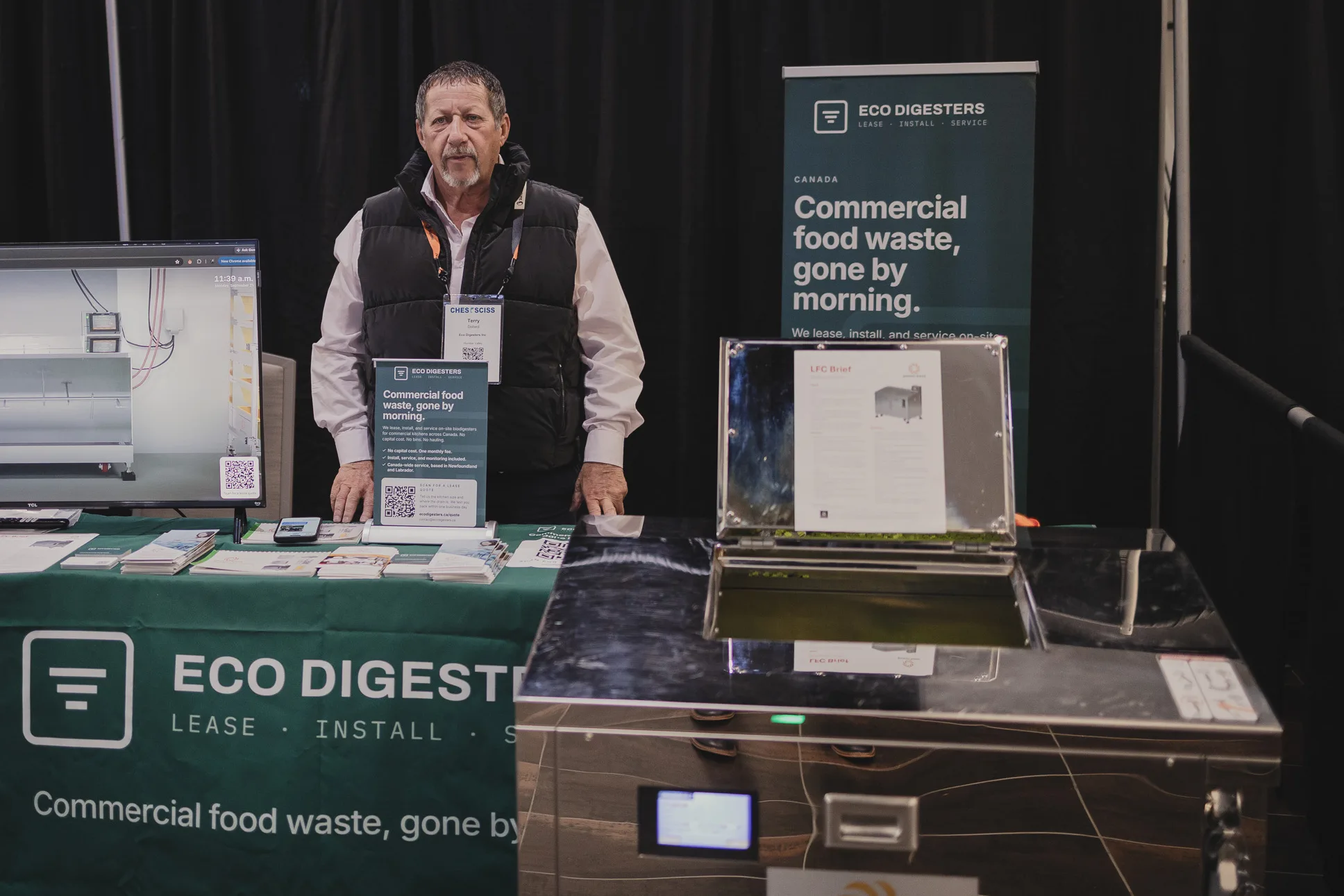 A member of our team behind the booth table, beside a stainless steel Eco Digester with its lid open, the booth screen, and a teal banner reading Commercial food waste, gone by morning.